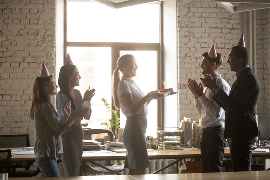 In a workspace, four adults wearing party hats clap for another, who holds a slice of cake bearing a lit candle