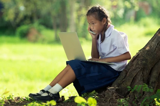 Girl sitting at the base of a tree on a laptop talking on the phone.