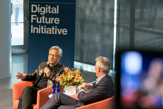 Two men sit on stage in front of a sign that says "Digital Future Initiative;" one speaks into a microphone